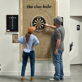 Two people with backs to the camera, one pointing at a dartboard mounted on a textured wall panel at The Clay Hole in Draper, Utah