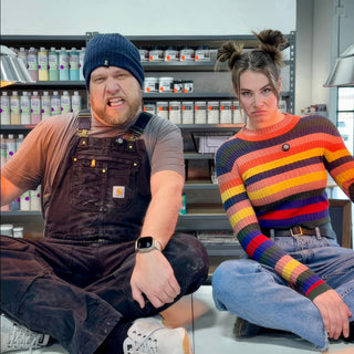 Two people sit cross-legged making stern faces in front of shelves of bottles and jars, one in dark overalls at The Clay Hole in Draper, Utah