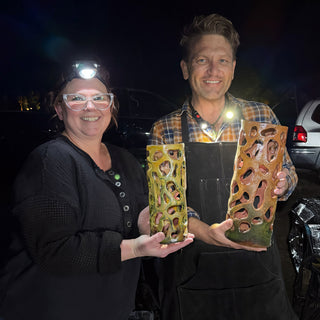 Two smiling people each hold a tall cylinder with cutouts, one yellow green and one amber, in the dark at The Clay Hole in Draper, Utah