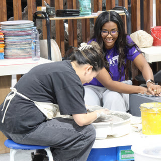 A potter bent over a pottery wheel with wet hands while a smiling person in a purple shirt leans in at The Clay Hole in Draper, Utah