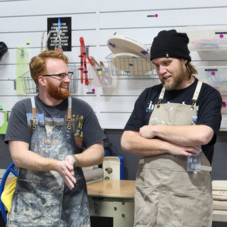Two potters in work aprons chatting in front of a wall of tools, one in a black beanie at The Clay Hole in Draper, Utah