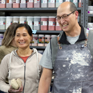 Two smiling potters stand in front of shelves of jars, one holding two lumps of clay at The Clay Hole in Draper, Utah