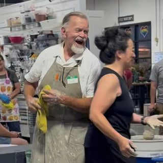 Two potters laugh together with clay on their hands at a pottery wheel, one holding a yellow cloth at The Clay Hole in Draper, Utah
