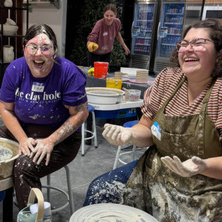 Two potters laugh with clay splattered on their faces, clothes and hands, seated near pottery wheels at The Clay Hole in Draper, Utah