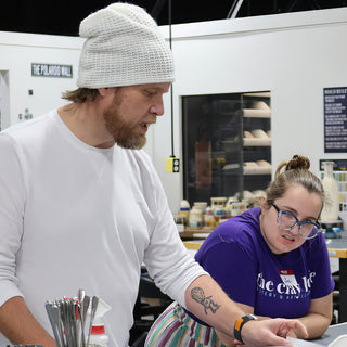 A potter in a white beanie leans over a worktable while a second potter in a purple shirt looks down at The Clay Hole in Draper, Utah