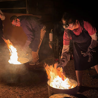Two people in safety goggles and gloves bend over separate burning metal cans in the dark, one wearing a headlamp at The Clay Hole in Draper, Utah