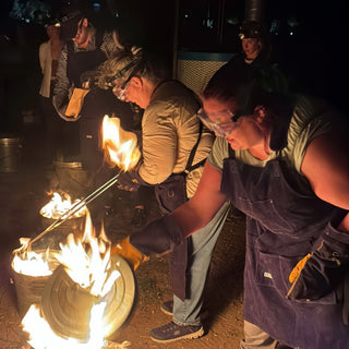 Two potters in goggles and denim aprons use long tongs and metal lids beside cans of open flames at The Clay Hole in Draper, Utah