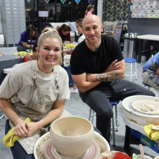 Two smiling potters pose behind a pale bowl on a pottery wheel, one with arms crossed at The Clay Hole in Draper, Utah