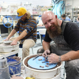 A bearded potter arranges small brown clay pieces on a blue disk as another potter shapes a bowl at The Clay Hole in Draper, Utah