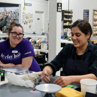 A potter in a purple shirt smiling at the camera beside another potter working a thin tool on clay at The Clay Hole in Draper, Utah