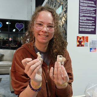 A smiling potter in glasses and a brown zip hoodie holding up two tiny unglazed clay figures in both hands at The Clay Hole in Draper, Utah