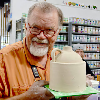 A smiling potter holding up a pale, unglazed clay jug with a loop handle on a green board at The Clay Hole in Draper, Utah
