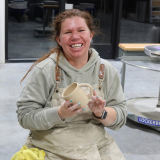 A potter in a tan apron grinning and holding up a small unglazed mug with a loop handle at The Clay Hole in Draper, Utah