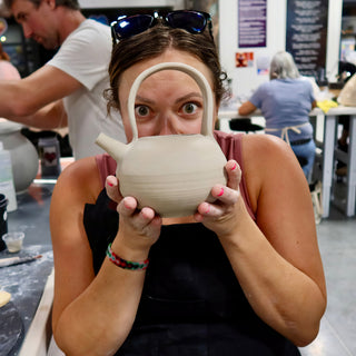 A potter peeks through the tall arched handle of a pale unglazed teapot with a short spout, eyes wide at The Clay Hole in Draper, Utah
