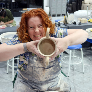 A potter in a clay-dusted apron laughing while holding up an unglazed two-handled cup with swirl marks inside at The Clay Hole in Draper, Utah