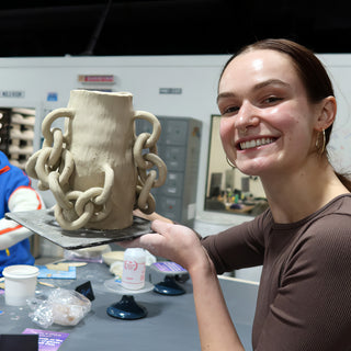 A smiling potter holding up an unglazed vase with large clay chain links draped down its sides at The Clay Hole in Draper, Utah