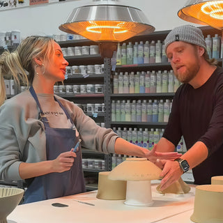 Two potters lean over a tan upside-down bowl on a small pedestal at a table, with shelves of bottles behind at The Clay Hole in Draper, Utah