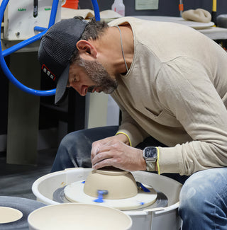 A potter in a black cap bending low over a pottery wheel to work on an upside-down cream bowl at The Clay Hole in Draper, Utah