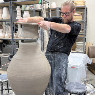 A potter holds a flat board with a gray clay form above the opening of a very tall unfired vase at The Clay Hole in Draper, Utah