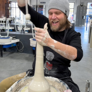 A laughing potter in a gray knit beanie stretching a very tall narrow clay vase upward on a pottery wheel at The Clay Hole in Draper, Utah