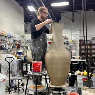 A potter in dark overalls reaching up to shape the rim of a very tall slender unglazed vase at The Clay Hole in Draper, Utah