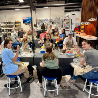 A class of potters at pottery wheels turn and wave at the camera from blue stools in a large room at The Clay Hole in Draper, Utah