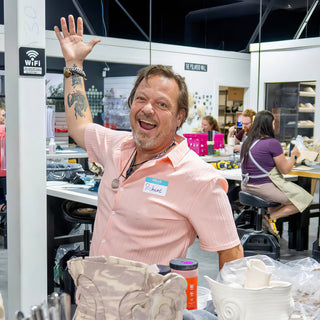 A grinning person in a pink shirt waving a tattooed arm above a worktable of pale clay pieces at The Clay Hole in Draper, Utah