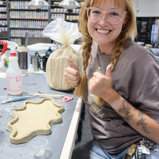 A smiling potter giving two thumbs up beside a flat clay dish with raised wavy edges at The Clay Hole in Draper, Utah