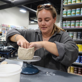 A potter pressing both hands around a thick, wavy-walled clay form sitting on a turntable at The Clay Hole in Draper, Utah