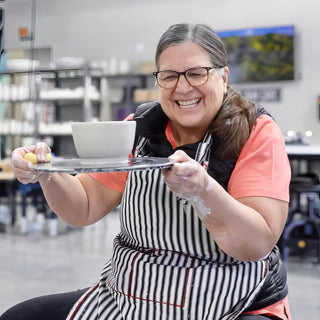 A laughing potter in a striped apron holds out a dark disk carrying a white bowl and a yellow sponge at The Clay Hole in Draper, Utah