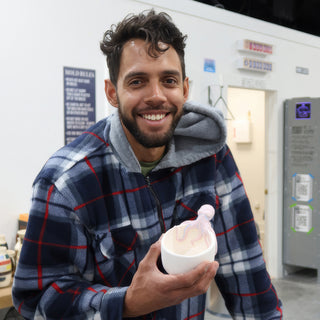 A potter in a plaid jacket smiling and holding a white bowl with a pink curled detail on its rim at The Clay Hole in Draper, Utah