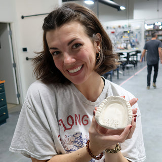 A smiling potter holding up a white ceramic dish shaped like a row of teeth with a stamped mark at The Clay Hole in Draper, Utah