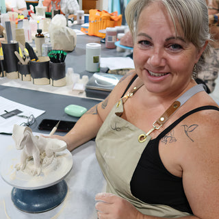 A smiling potter with a thin tool sits beside a white clay creature sculpture on a blue turntable at The Clay Hole in Draper, Utah