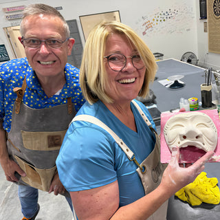 Two smiling potters pose together while one holds up a white clay face mask on a pink board at The Clay Hole in Draper, Utah