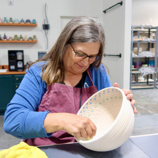 A potter wiping the inside of a white bowl with ridged sides and tiny blue and green dots at The Clay Hole in Draper, Utah