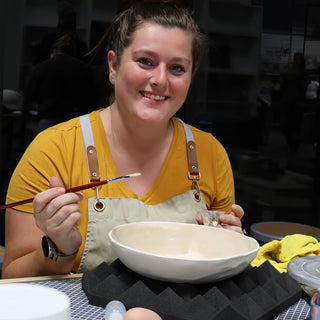 A potter in a mustard shirt holding a thin brush over a wide cream bowl with a wavy rim at The Clay Hole in Draper, Utah