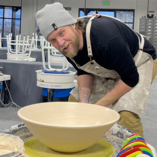 A potter leaning in behind a wide, smooth clay bowl with a flared rim sitting on a pottery wheel at The Clay Hole in Draper, Utah