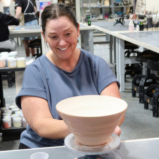 A potter smiling down at a wide pale pink bowl with horizontal ridges sitting on a turntable at The Clay Hole in Draper, Utah