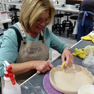 A smiling potter works a yellow sponge inside a wide dish with a ruffled rim on a purple board at The Clay Hole in Draper, Utah