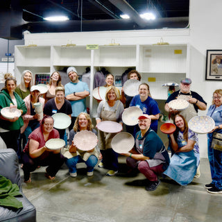A group of members crowded together in two rows, holding up painted plates and bowls in soft pastel colors at The Clay Hole in Draper, Utah