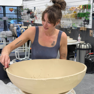 A potter works the rim of a very large tan clay bowl with a small tool at The Clay Hole in Draper, Utah