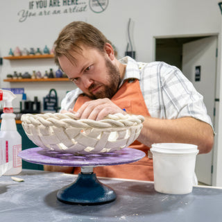 A potter leans over a white woven clay basket with a braided rim, resting on a purple turntable at The Clay Hole in Draper, Utah