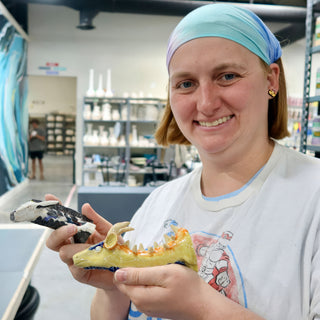 A potter in a headband holds a yellow glazed dragon figure with spiky teeth and a black and white piece at The Clay Hole in Draper, Utah