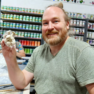 A smiling potter holds up a small cluster of cream clay mushrooms in front of shelves of jars during a raku firing with The Clay Hole, a pottery studio in Draper, Utah