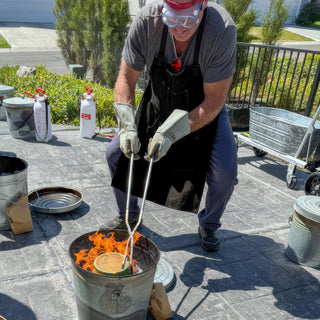A potter in goggles and a black apron grips a small piece with long tongs over a bucket of fire during a raku firing with The Clay Hole, a pottery studio in Draper, Utah