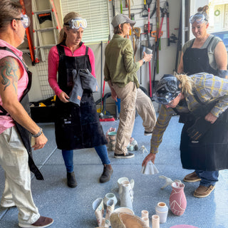 A group in aprons and goggles gathers around white and pink vases set out on a garage floor during a raku firing with The Clay Hole, a pottery studio in Draper, Utah