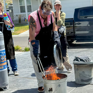 A person in a pink shirt and black apron holds a metal lid and long tongs over flames during a raku firing with The Clay Hole, a pottery studio in Draper, Utah