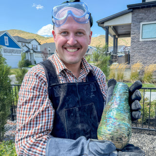 A smiling potter with goggles on their head holds a teardrop shaped vase with ribbed iridescent blue and gold glaze during a raku firing with The Clay Hole, a pottery studio in Draper, Utah