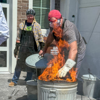 A person in a red cap and goggles lifts the lid off a metal can as tall flames rise during a raku firing with The Clay Hole, a pottery studio in Draper, Utah
