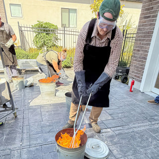 A potter in a green cap, goggles and gloves lowers long tongs into a metal bucket of flames during a raku firing with The Clay Hole, a pottery studio in Draper, Utah
